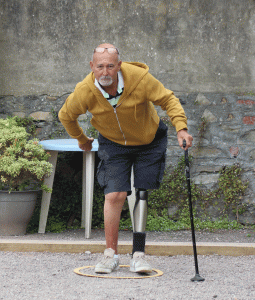 This image shows a male player with a prosthetic left leg, with a supporting stick, standing in the playing circle preparing to throw his boule.